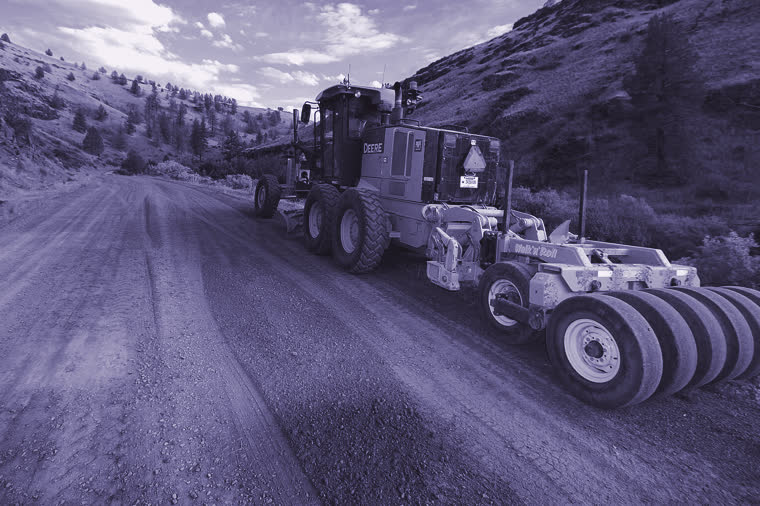 A road grader and a towed roller parked on a graded dirt road in dry hill country.